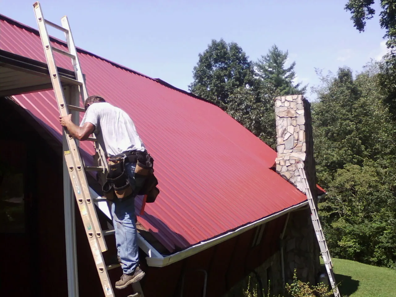 Roofer on a ladder beside a red metal roof and stone chimney — Soffit & Fascia Repair by Roofing Brothers in Grovetown